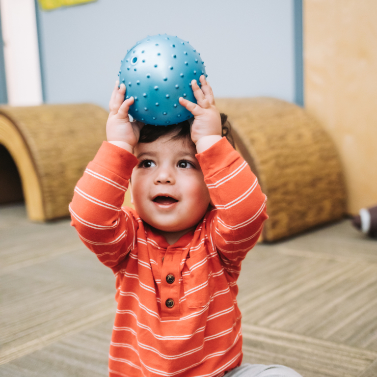 A young child wearing an orange and white tshirt holding a blue bouncy ball over his head