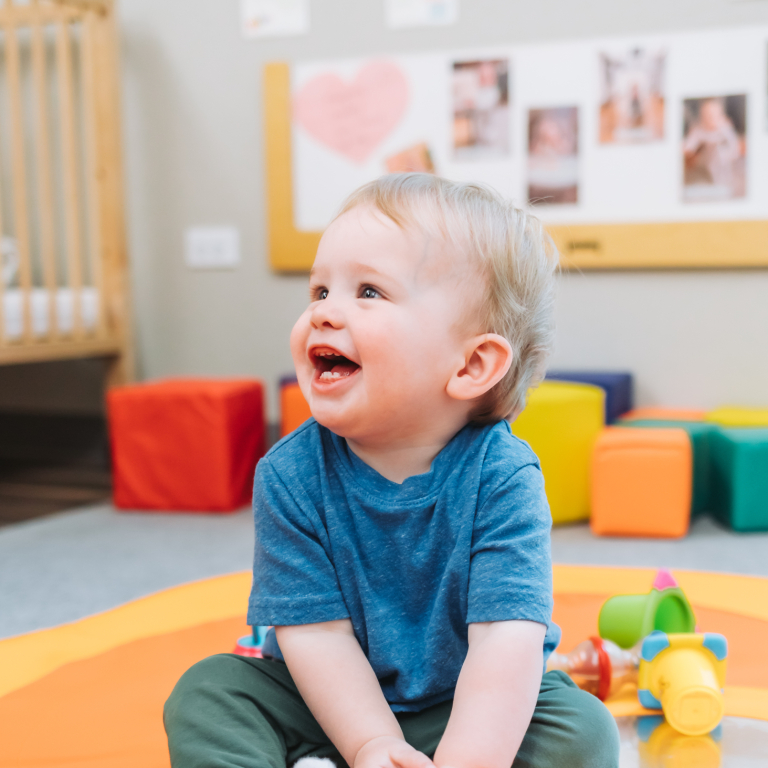 A young boy sat on a play mat surrounded by toys smiling