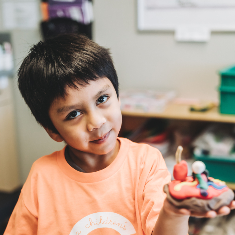 A young boy with dark hair holding a handmade craft out in one hand