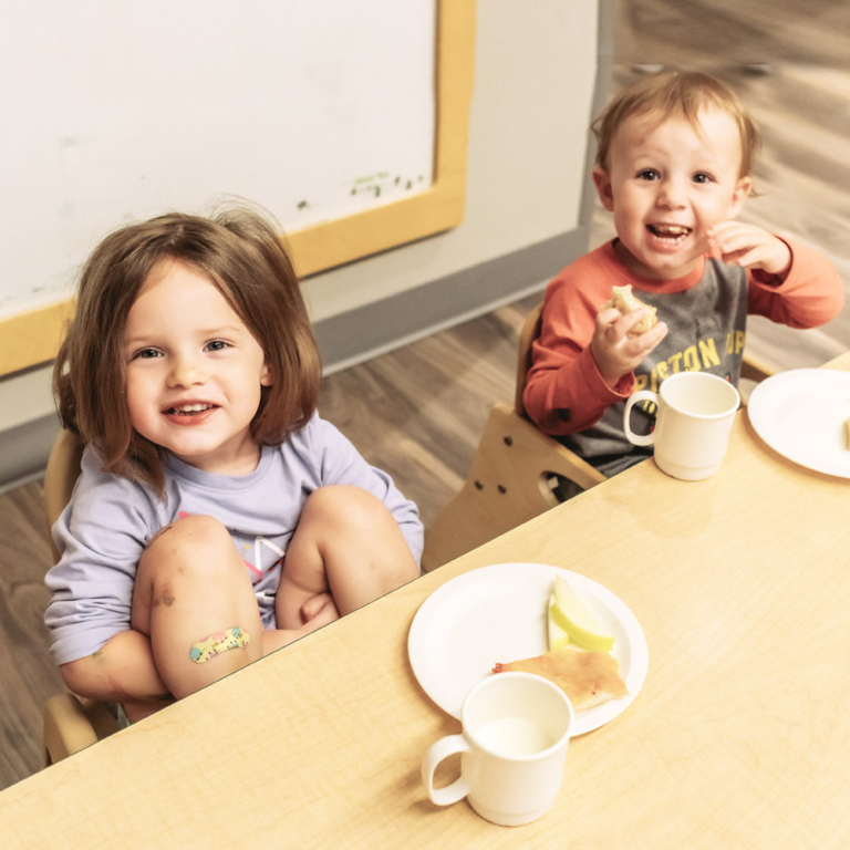 Two young children sat at a table eating food off a plate