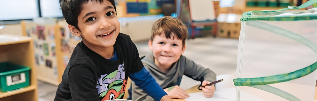 Two young boys sat at a desk smiling into the camera